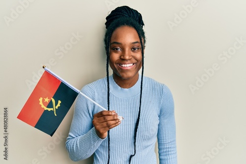 African american woman with braided hair holding angola flag looking positive and happy standing and smiling with a confident smile showing teeth photo