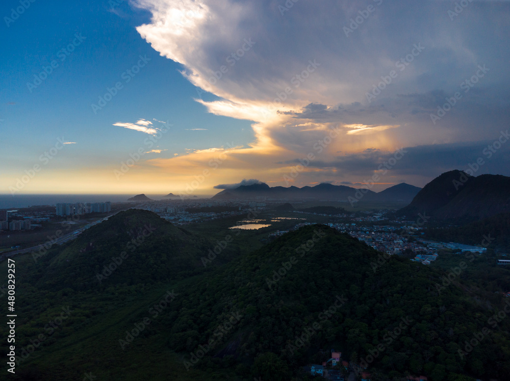 Obraz premium Hills in Rio de Janeiro, Brazil with rain clouds forming at sunset. Aerial view of Ilha Pura Park. In the background the beach of Recreio dos Bandeirantes. Sunny Day. Drone take