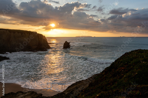 Portuguese sunset landscape with the sun's rays through the clouds on the shores of the Atlantic Ocean with sheer high cliffs and foamy waves. A small island is visible on the horizon. Peniche