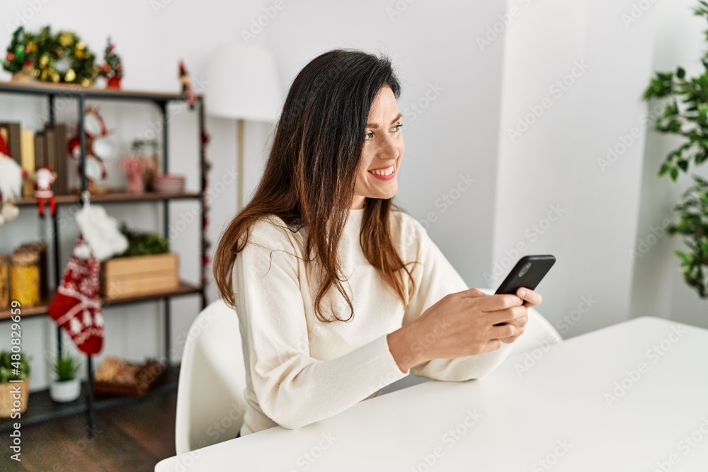 Middle age hispanic woman using smartphone sitting on the table by christmas decor at home Middle age hispanic woman using smartphone sitting on the table by christmas decor at home