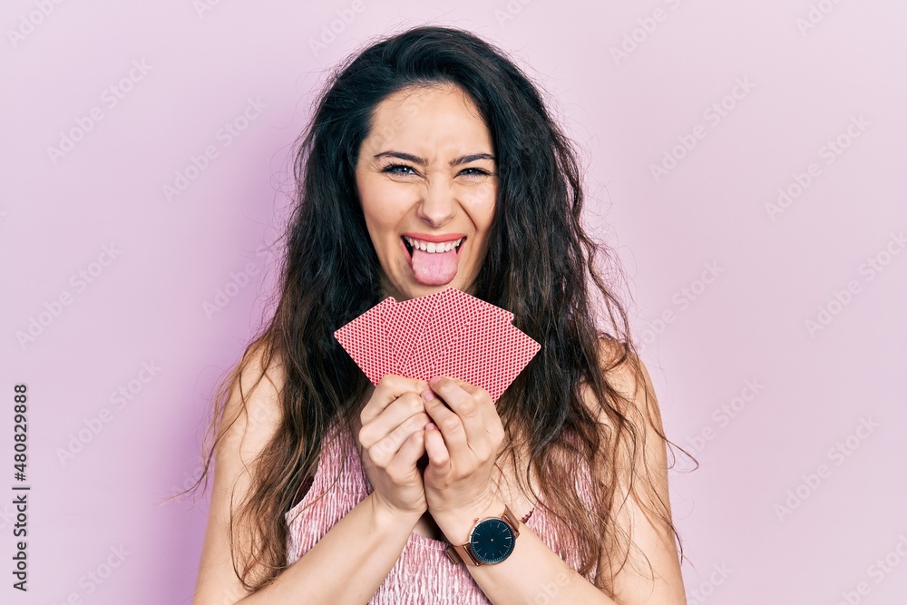 Young hispanic woman covering mouth with cards sticking tongue out happy with funny expression. Young hispanic woman covering mouth with cards sticking tongue out happy with funny expression.