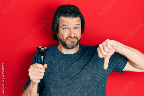 Middle age caucasian man holding electric razor machine with angry face, negative sign showing dislike with thumbs down, rejection concept