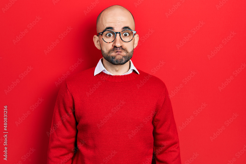 Young bald man wearing casual clothes and glasses puffing cheeks with funny face. mouth inflated with air, crazy expression. Young bald man wearing casual clothes and glasses puffing cheeks with funny face. mouth inflated with air, crazy expression.