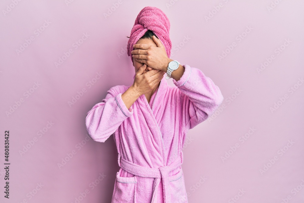 Young blonde woman wearing shower towel cap and bathrobe covering eyes and mouth with hands, surprised and shocked. hiding emotion Young blonde woman wearing shower towel cap and bathrobe covering eyes and mouth with hands, surprised and shocked. hiding emotion
