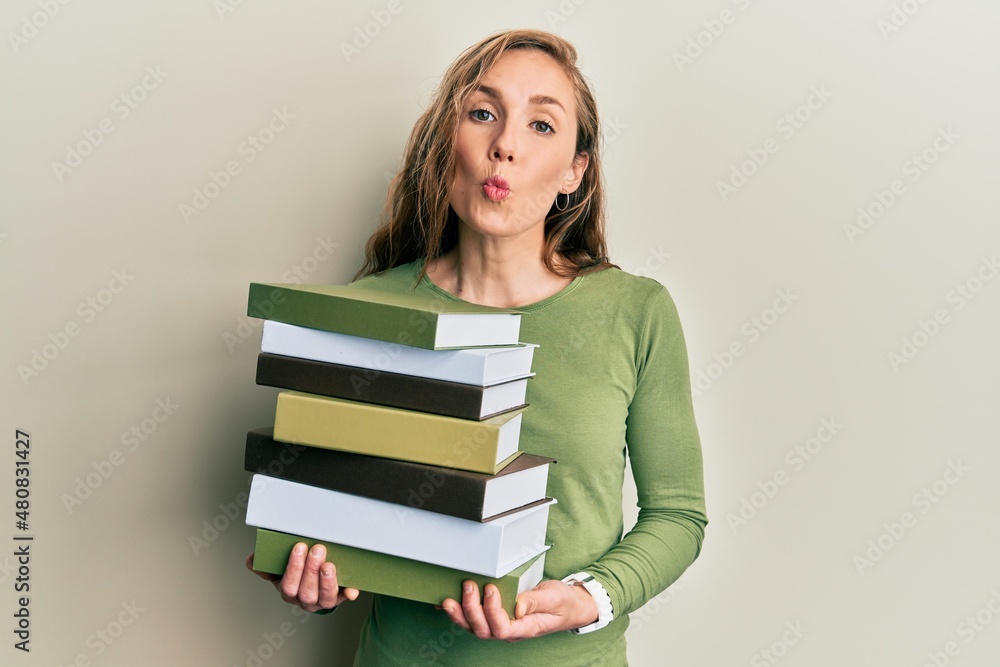 Young blonde woman holding a pile of books looking at the camera blowing a kiss being lovely and sexy. love expression. Young blonde woman holding a pile of books looking at the camera blowing a kiss being lovely and sexy. love expression.