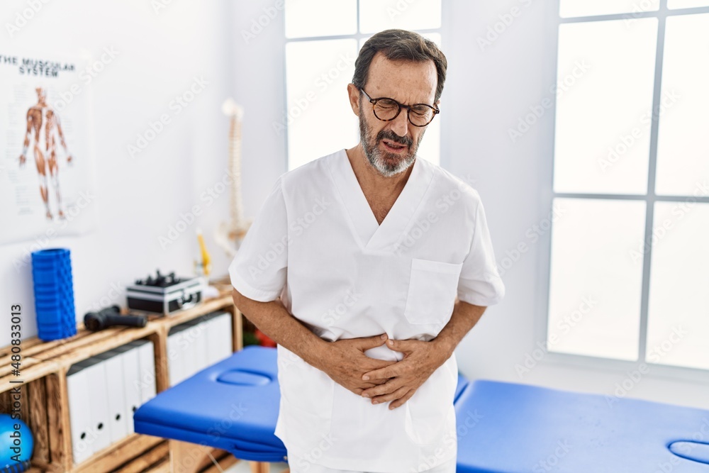 Middle age man with beard working at pain recovery clinic with hand on stomach because indigestion, painful illness feeling unwell. ache concept. Middle age man with beard working at pain recovery clinic with hand on stomach because indigestion, painful illness feeling unwell. ache concept.