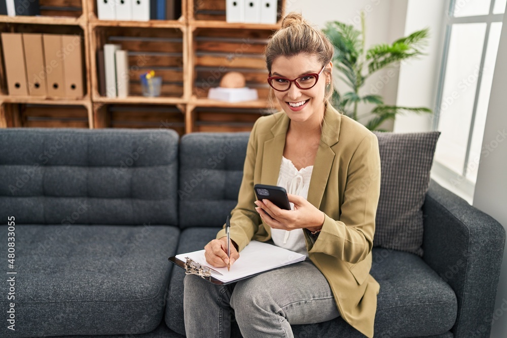 Obraz premium Young blonde woman psychologist using smartphone sitting on sofa at psychology center