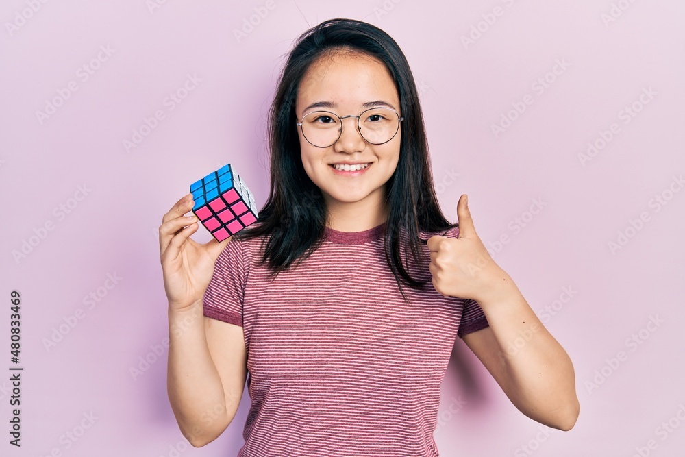 Young chinese girl playing colorful puzzle cube intelligence game smiling happy and positive, thumb up doing excellent and approval sign Young chinese girl playing colorful puzzle cube intelligence game smiling happy and positive, thumb up doing excellent and approval sign