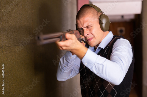 Focused man in ear protectors aiming single barrelled shotgun at target in shooting range.