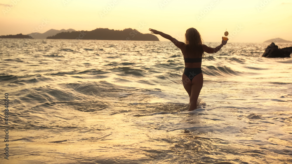 Woman stand knee-deep in sea on sunset. Girl on beach in green swimsuit having fun and waving his hands, drinks her orange cocktail