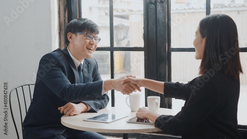 Happy two Asian young businessman and woman shaking hands greeting before meeting or negotiation with digital tablet sitting on desk cafe, Businesspeople discussion planning and smile lunch