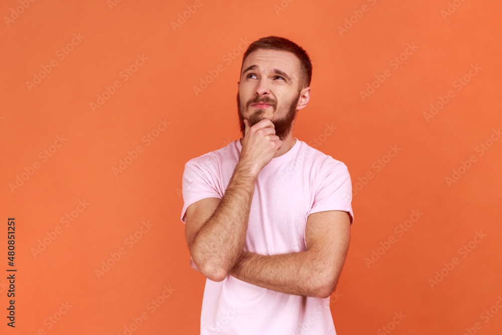Portrait of thoughtful handsome bearded man holding his chin and pondering idea, confused not sure about solution, wearing pink T-shirt. Indoor studio shot isolated on orange background. Portrait of thoughtful handsome bearded man holding his chin and pondering idea, confused not sure about solution, wearing pink T-shirt. Indoor studio shot isolated on orange background.