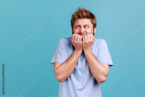 Anxiety disorder. Portrait of stressed out, worried bearded man biting nails, nervous about troubles, panicking and looking scared. Indoor studio shot isolated on blue background. photo