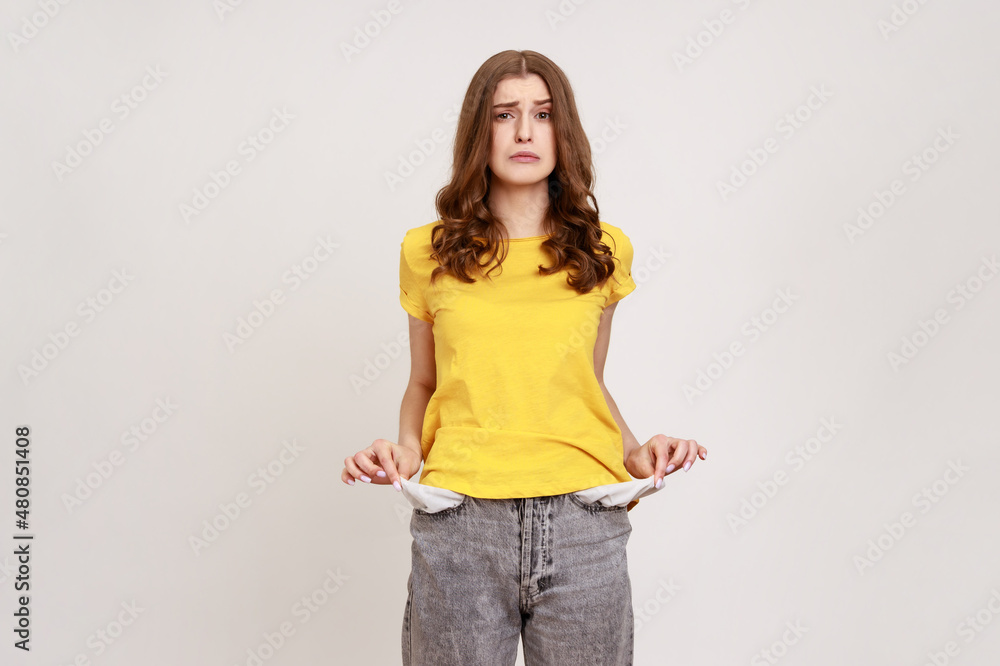 Portrait of frustrated worried brown haired female of young age in casual yellow T-shirt turning out empty pockets showing no money gesture. Indoor studio shot isolated on gray background. Portrait of frustrated worried brown haired female of young age in casual yellow T-shirt turning out empty pockets showing no money gesture. Indoor studio shot isolated on gray background.