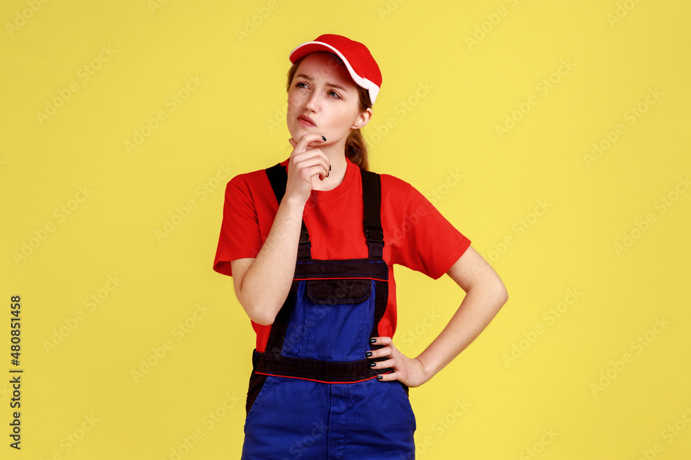 Portrait of pensive handy woman standing and holding chin, looking away with thoughtful facial expression, wearing overalls and red cap. Indoor studio shot isolated on yellow background.