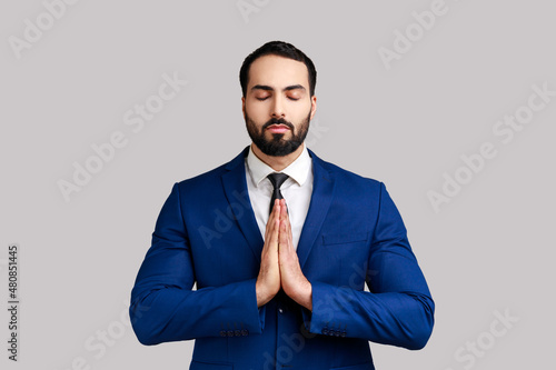 Portrait of calm handsome bearded businessman standing in yoga pose and try to relaxing, keeps palms together, wearing official style suit. Indoor studio shot isolated on gray background. photo