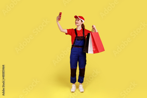 Full length portrait of courier woman standing with shopping bag in hands and taking selfie or having video call with client, wearing overalls and cap. Indoor studio shot isolated on yellow background