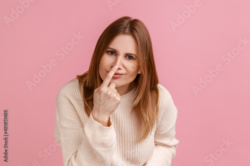 Portrait of attractive young blond woman pointing at her nose, saying to trickster you are liar, body language, wearing white sweater. Indoor studio shot isolated on pink background. photo