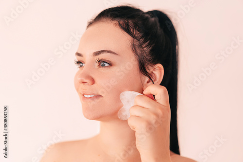 a beautiful brunette girl massages with a gouache scraper. a young woman of European appearance with clean, fresh skin. Jade roller facial massager made of natural stone. Cosmetology