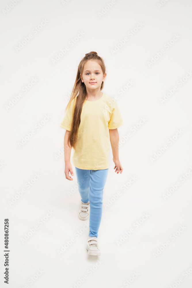 A child in a yellow T-shirt and blue pants. Baby with long blonde hair. Beautiful girl with a model appearance on a light background A child in a yellow T-shirt and blue pants. Baby with long blonde hair. Beautiful girl with a model appearance on a light background