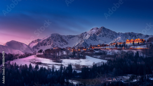 Scenic view of a town with hotels against blue sky and snowcapped mountains at sunset in winter. Snow and forest in the foreground