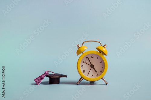 Yellow alarm clock with graduation cap on blue background closeup with copy space
