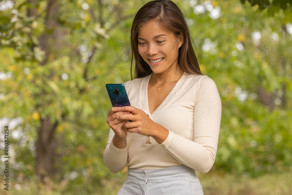 Asian woman using mobile phone outside walking in summer park commuting to work. Happy bipoc young businesswoman smiling holding device. Asian woman using mobile phone outside walking in summer park commuting to work. Happy bipoc young businesswoman smiling holding device.