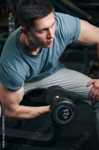 Concentrated sportsman lifting one dumbbell from seated position