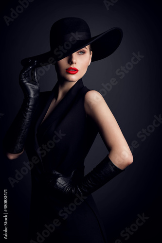 Fashion Model in Wide Brim Hat with Red Lips Make up. Elegant Retro Woman in Black Suit Dress and Gloves over Dark Gray Studio Background
