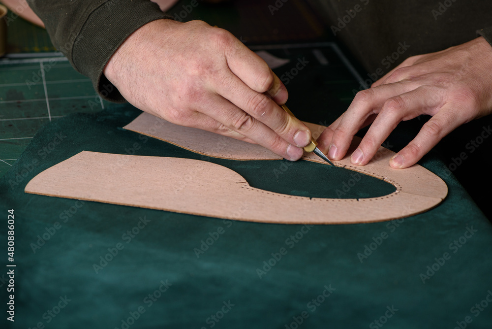 Shoemaker hands holding craft knife cutting green suede leather for handmade shoes on his workplace in workshop. Closeup. Copy space.