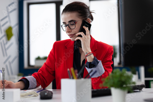 Businesswoman taking notes in smartphone phonecall sitting at desk. Entrepreneur with glasses working in startup office. Small business owner in red jacket writing on paper in startup enviroment.