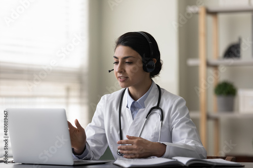 Happy young female Indian family doctor therapist in headphones with mic consulting patient online by computer video call conversation, telling advices or taking part in educational virtual event.