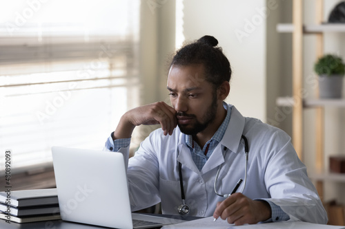 Anxious unhappy young male African American general practitioner doctor therapist in white uniform looking at computer screen, feeling stressed thinking of difficult medical problem solution in clinic