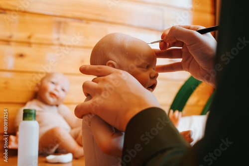Foto Close-up female hands of craftsman maker decorating with brush realistic newborn caucasian baby doll, painting hair on head of reborn sitting at workplace, indoors