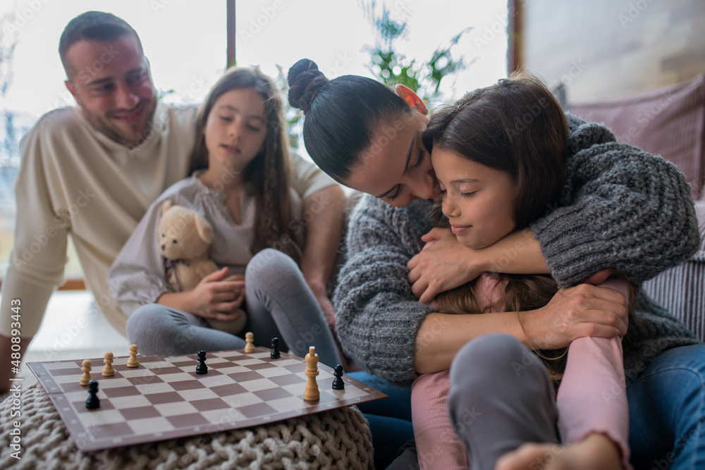 Two happy sisters with mother and father sitting on floor and playing chess together at home