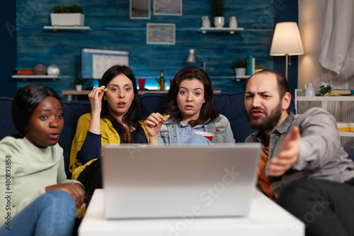 Portrait of multi-ethnic friends sitting on couch watching online movie on laptop computer enjoying hanging out together. Group of people enjoying relaxing time in living room. Friendship concept