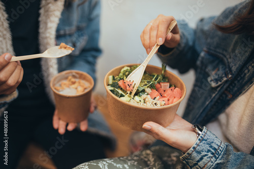 Two young women female friends sitting outdoors eating takeaway food, laughing and having fun. Food delivery and takeout.