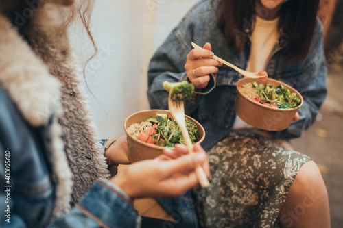 Photos Two young women female friends sitting outdoors eating takeaway food, laughing and having fun