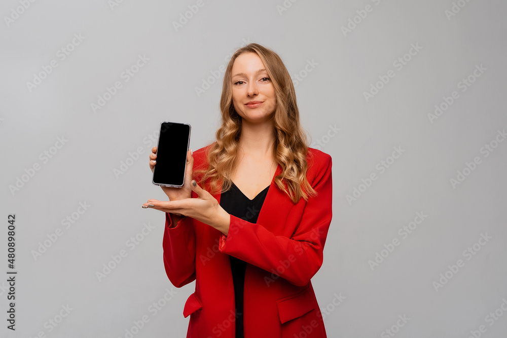 Naklejka premium beautiful and confident business woman showing smartphone screen, demonstrating mobile app, standing in red blazer against gray background