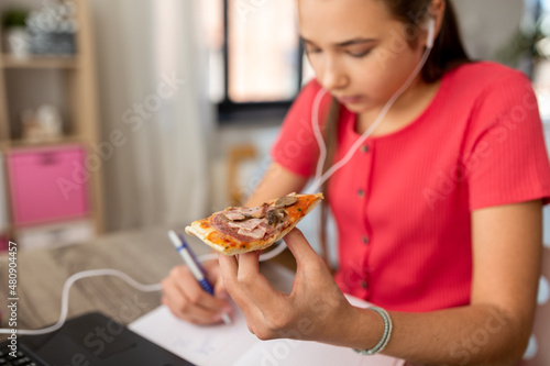 children, education and distant learning concept - close up of teenage student girl in earphones with laptop computer eating pizza and writing to notebook at home