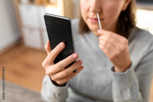 medicine, quarantine and pandemic concept - close up of woman with swab and smartphone taking sample from her nose and making nasal coronavirus self test at home
