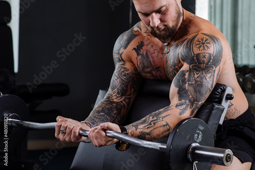 Muscular tattooed athlete swings his biceps with a barbell in the gym