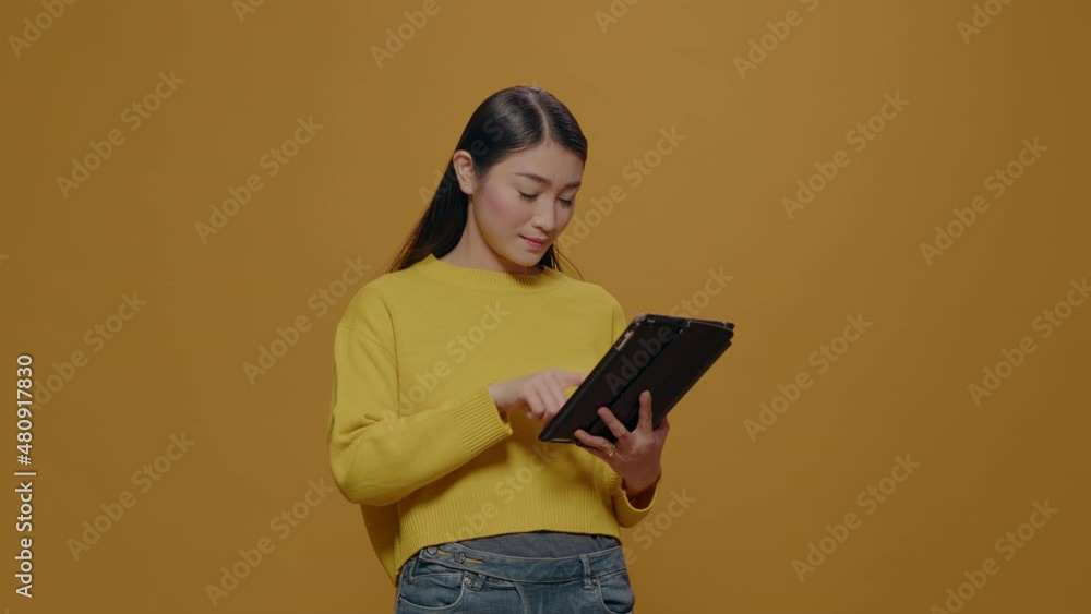 Focused model browsing internet on digital tablet in studio, using device with touch screen to do successful work. Asian woman holding modern gadget with app connection over background. Focused model browsing internet on digital tablet in studio, using device with touch screen to do successful work. Asian woman holding modern gadget with app connection over background.