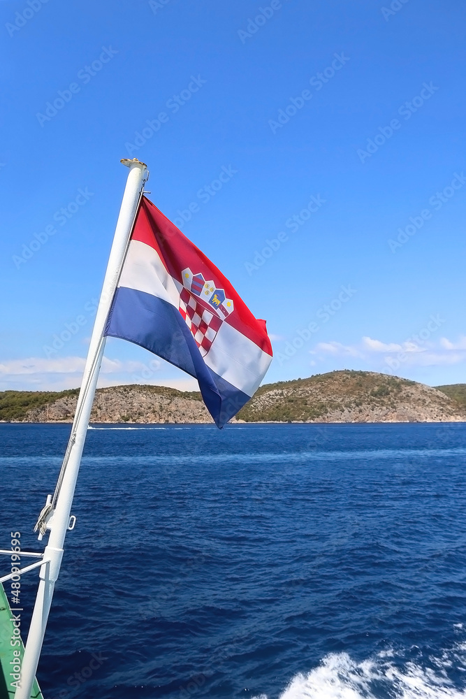 Fototapeta premium Flag of Croatia blowing in the wind. Adriatic sea, island and boats in the background.