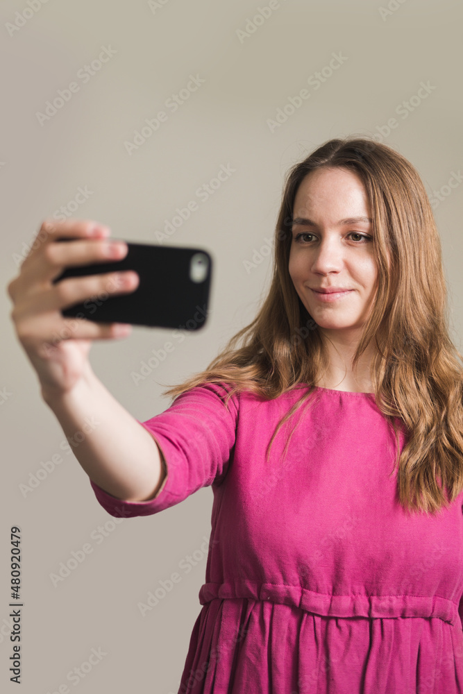 Naklejka premium Young millennial woman in pink dress with long hair makes a selfie at home.