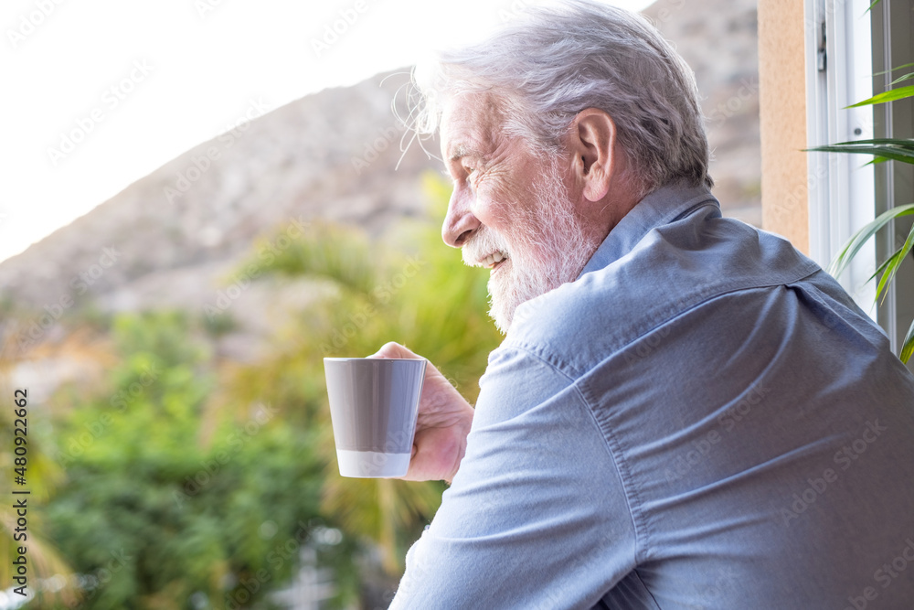 Smiling attractive adult mature senior man standing at the window holding a coffee cup of tea looking away. Elderly white haired bearded grandfather in relaxed moment having a break Copy space Smiling attractive adult mature senior man standing at the window holding a coffee cup of tea looking away. Elderly white haired bearded grandfather in relaxed moment having a break Copy space
