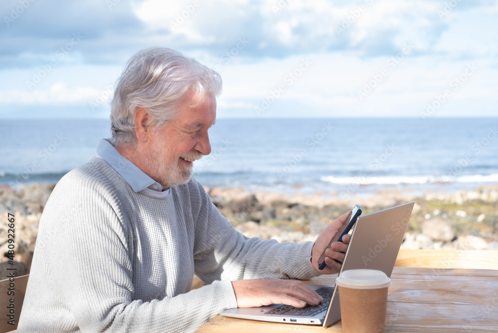 Smiling attractive mature senior adult professional business man in remote working on laptop computer by the beach. Elderly bearded male holding smartphone sitting at a wooden table close to the sea