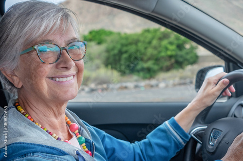 Happy active mature adult senior woman granny driving the car, looking at camera smiling. Elderly female wearing glasses holding wheel enjoying travel and freedom with her automobile
