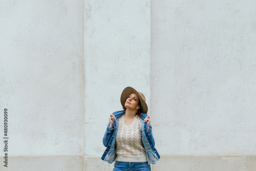 An adult woman in a denim jacket and beige hat posing against a wall while looking up