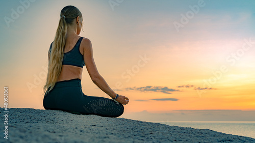 Young caucasian woman with long hair meditating in a black clothes for yoga on a rocks on the top by the sea. On the background colorful sunrise sky.